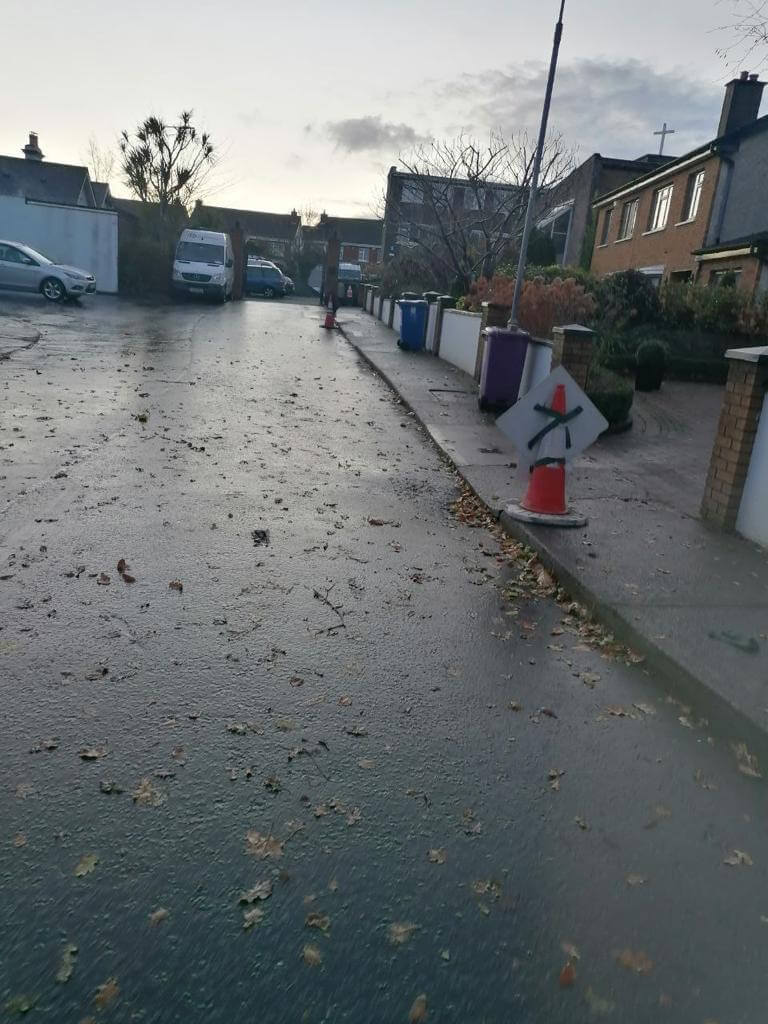 Wet freshly laid tarmac with houses, cars, trash bins and traffic cone
